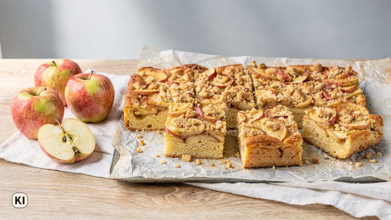 KI-Bild: Apfelkuchen vom Blech mit Apfelscheiben und Zimtstreuseln, drei Stücke herausgenommen, links ganze und ein halbierter Apfel auf Leinen
