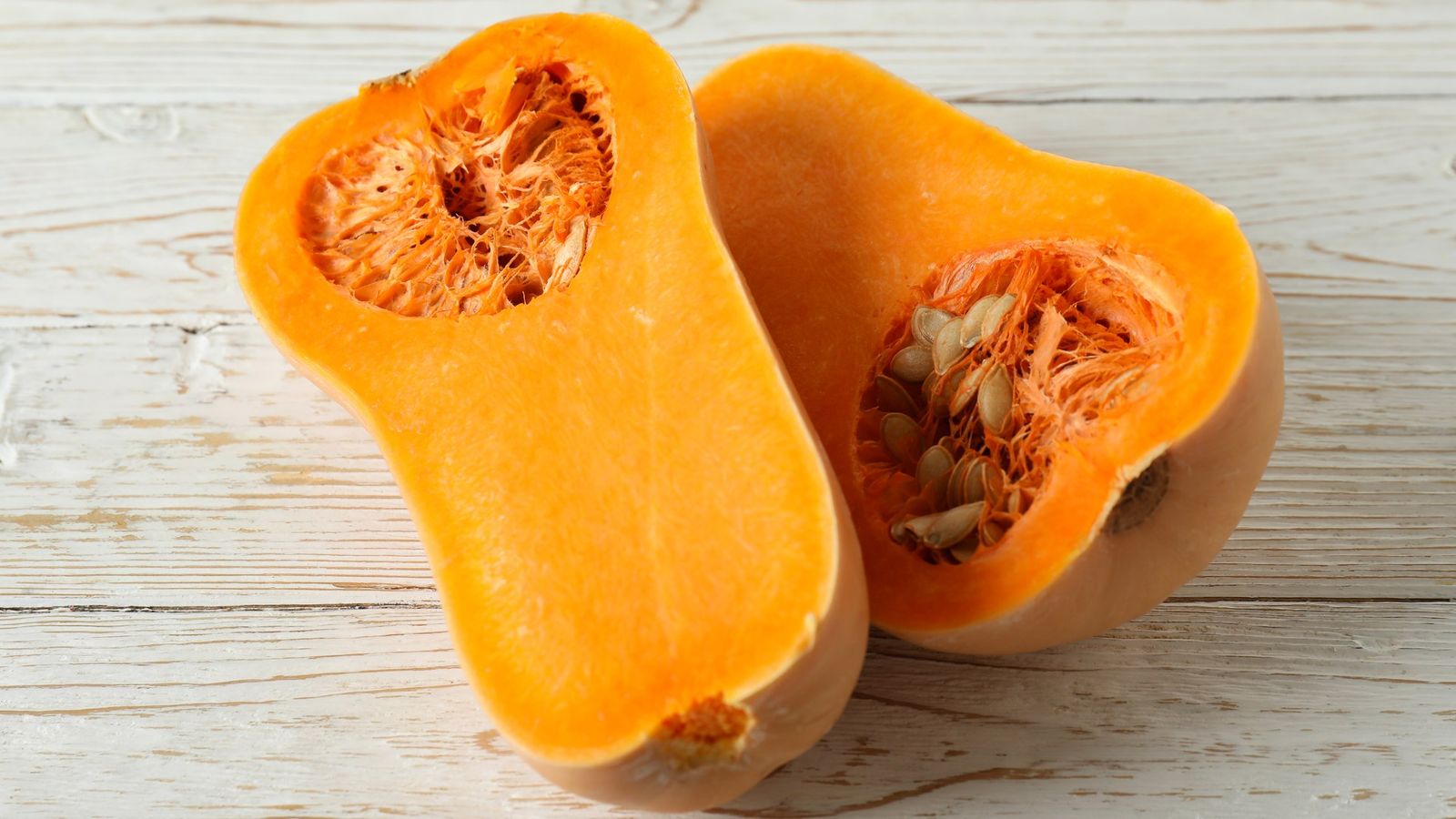 Ein längs halbierter Butternut-Kürbis mit orangefarbenem Fruchtfleisch, Kernen und heller Schale auf weiß gestrichenem Holz