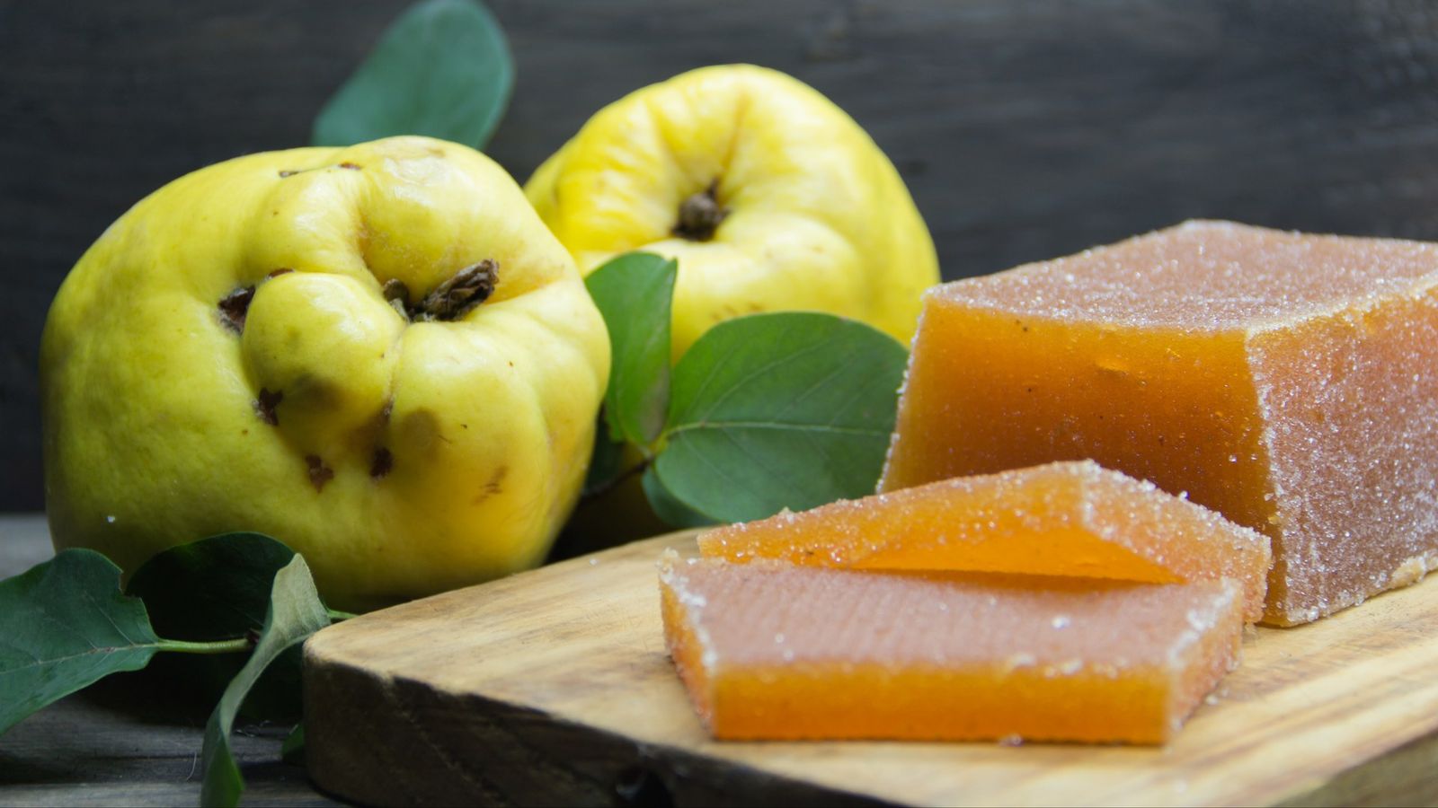 Quittenbrot in Rauten geschnitten und in Zucker gewendet auf einem Holzbrett, dahinter zwei gelbe Quitten mit grünen Blättern
