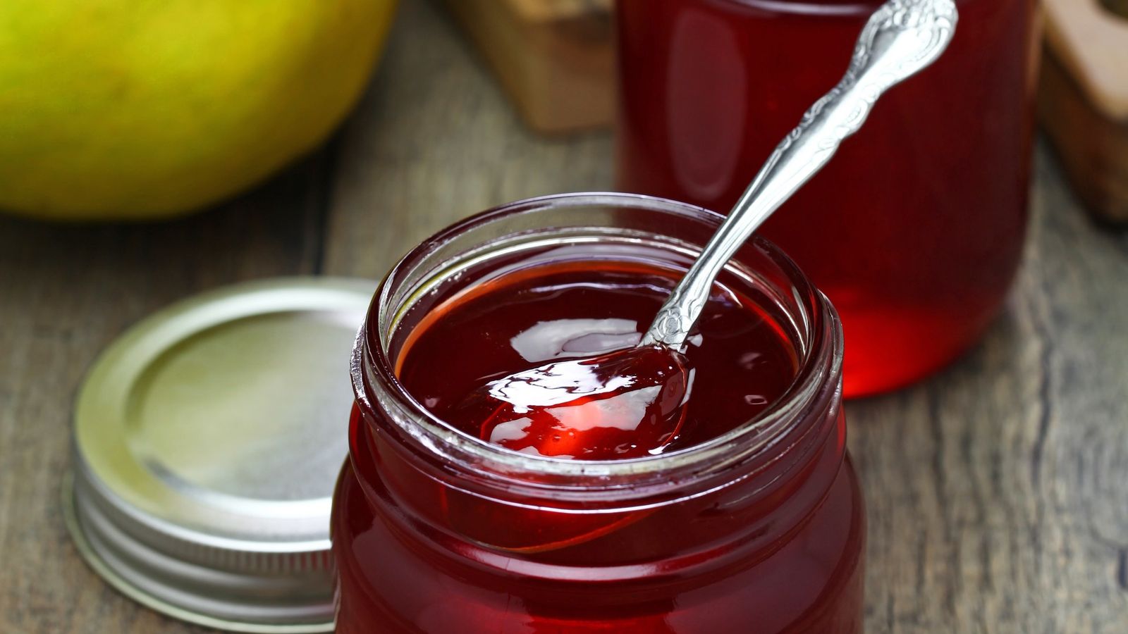 Rotes Quittengelee in einem offenen Glas mit Löffel, dahinter ein zweites Glas und gelbgrüne Quitten auf einem Holztisch, der Deckel liegt daneben